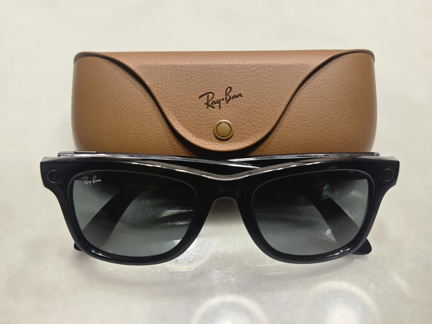 검은색 Ray-Ban Meta Gen 1 스마트 글래스 한 쌍이 갈색 가죽 충전 케이스 앞에 놓여 있는 사진.